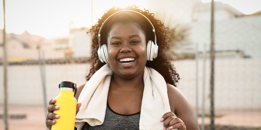 image of black woman wearing active gear, holding a yellow water bottle with headphones on and a towel around her neck