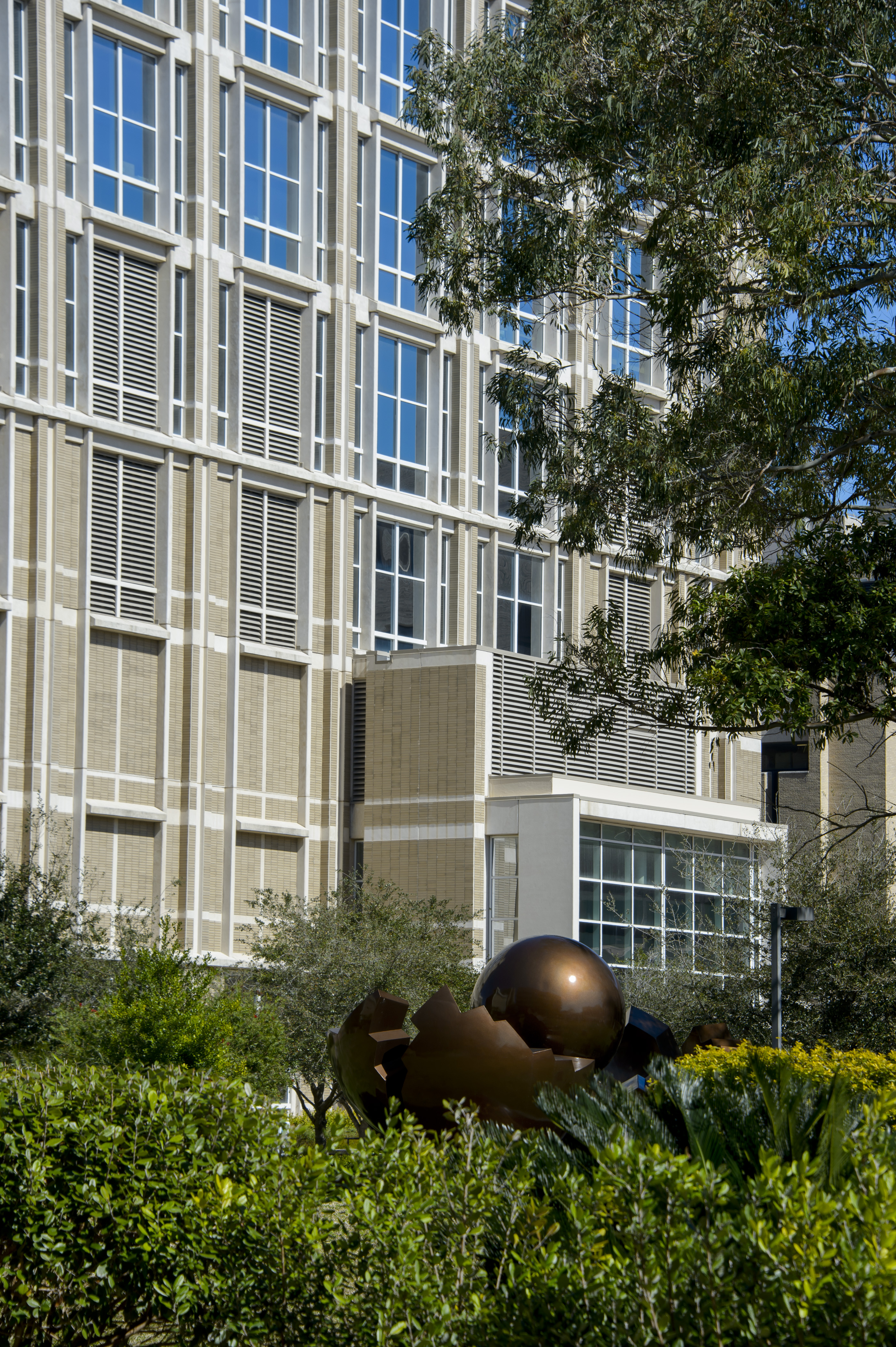 Exterior view of a UTMB campus building with modern windows, landscaped greenery, and an abstract metal sculpture in the foreground.