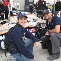 Two people pose beside an IndyCar race car in the pit area.
