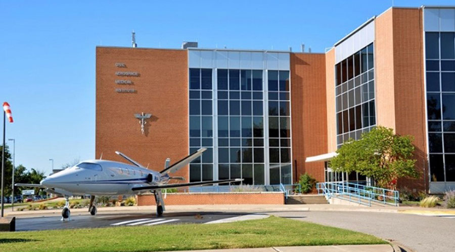 Large brick and glass L-shaped building with circular drive and static display of aircraft at FAA Civil Aerospace Medical Institute in Oklahoma City.