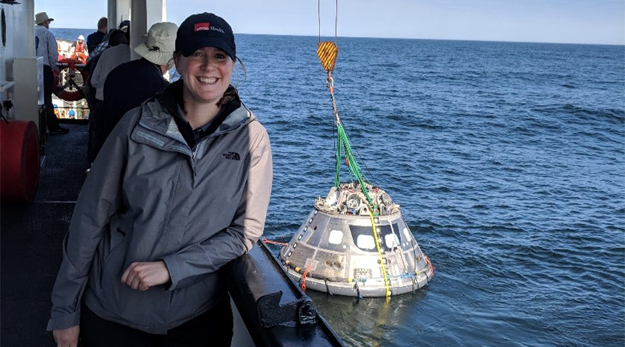 UTMB Aerospace Medicine Resident on a ship, with the Orion capsule in the water attached to a crane in Galveston Bay behind her. The resident was supporting medical operations testing of the Orion capsule in advance of the Artemis campaign.