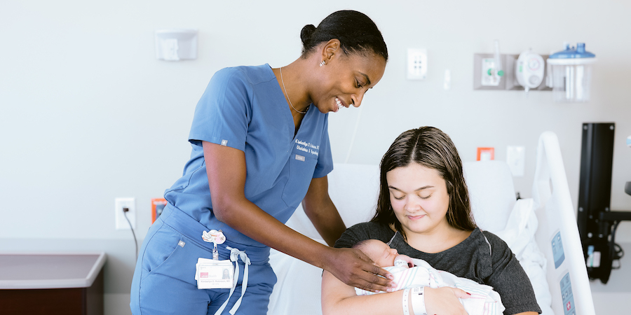 Health care provider wearing blue scrubs stands beside a seated adult holding a swaddled newborn in a hospital room, with medical equipment and a hospital bed visible in the background, illustrating newborn care and a pediatric visit
