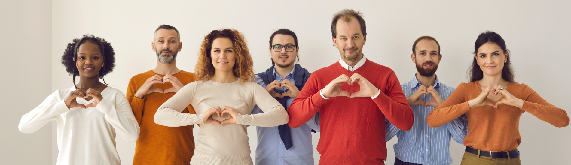 A diverse group of adults standing side by side, each forming a heart shape with both hands at chest level against a neutral background.