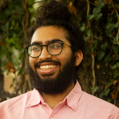Individual wearing glasses and a button‑down shirt standing in front of dense green foliage.