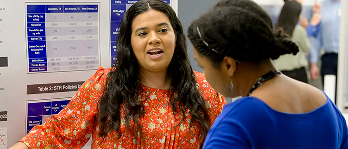 Two individuals stand facing each other and speak in front of a research poster displaying tables and charts during an academic event at the University of Texas Medical Branch.