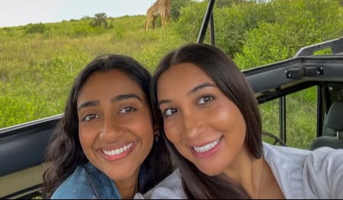 Two UTMB medical students, Nidhi Subramanian and Zeyanna Dhalla, smiling inside an open safari vehicle with a giraffe standing in the grassy landscape behind them in Nairobi, Kenya.