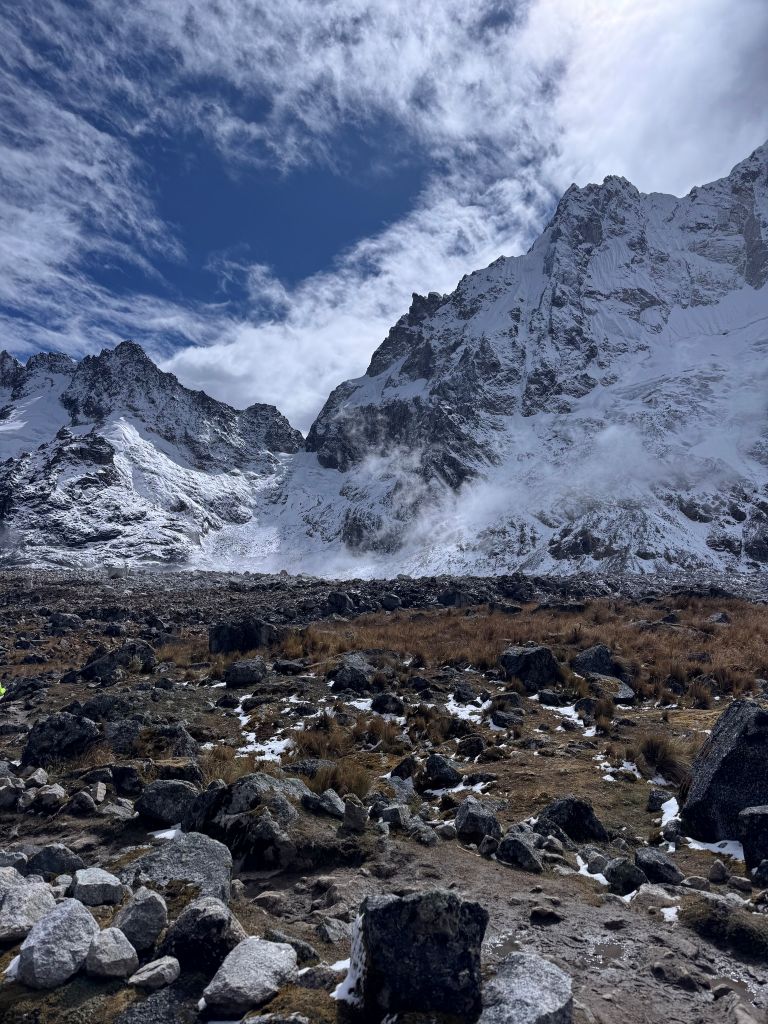 Fowler_7 Snow-dusted mountains under a partly cloudy sky, with rocky terrain and patches of grass in the foreground.