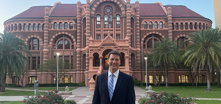 Individual standing on the University of Texas Medical Branch campus in front of a historic academic building with arched architecture and palm trees.