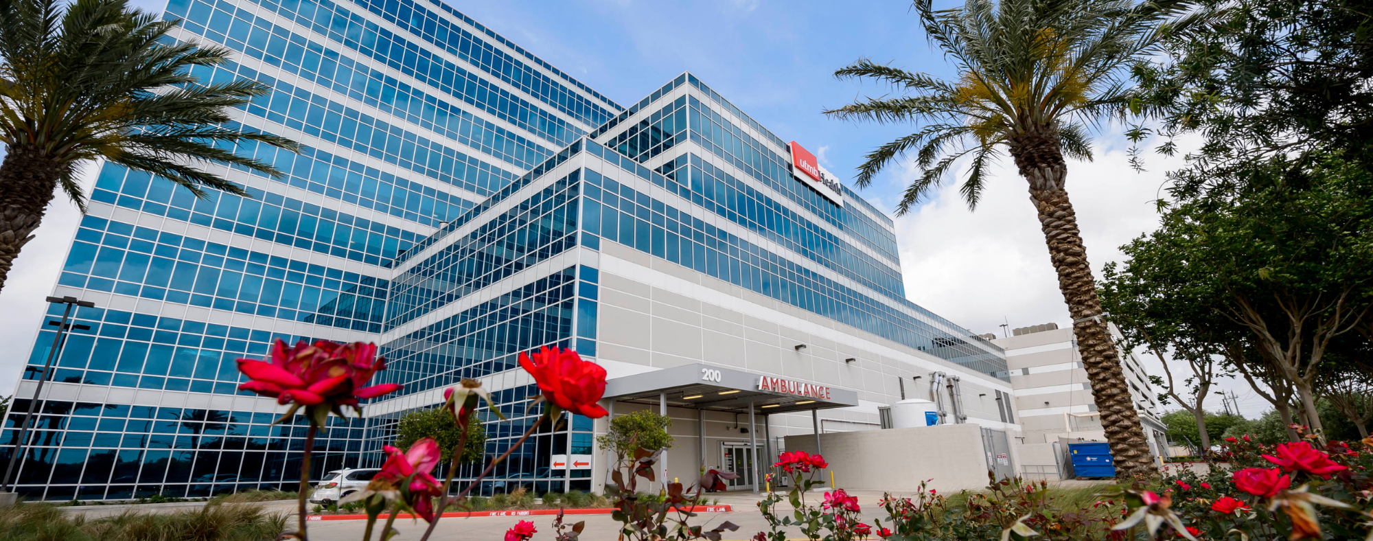 Exterior view of a modern UTMB Health hospital building with glass façade, palm trees, and ambulance entrance canopy surrounded by red flowers.