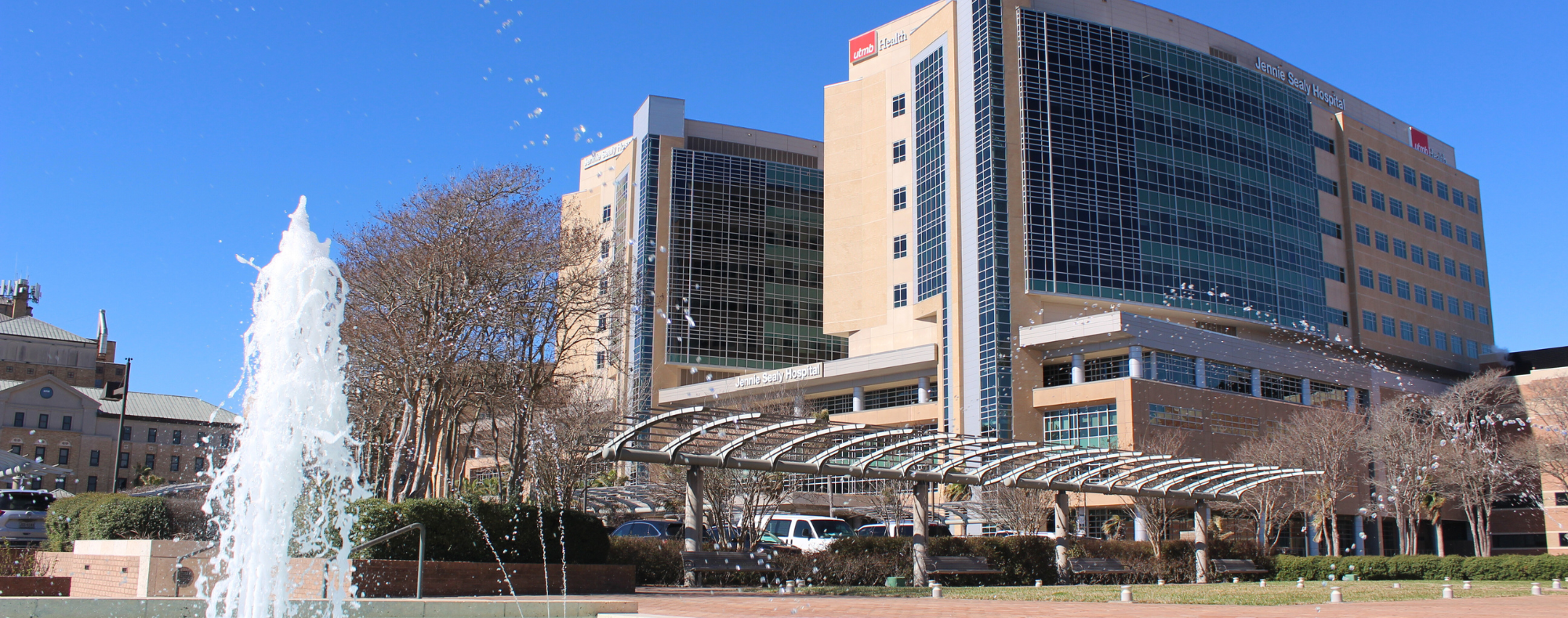 Exterior view of UTMB Health Jennie Sealy Hospital with a modern glass façade, fountain in the foreground, and covered walkway leading to the entrance.