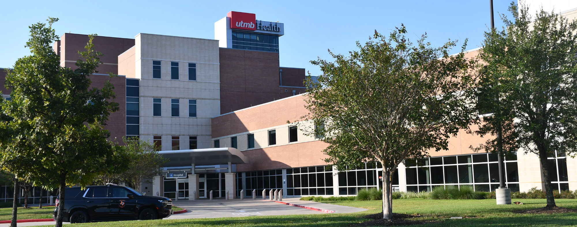 Exterior view of a UTMB Health hospital building with brick and glass façade, main entrance canopy, and trees lining the driveway.