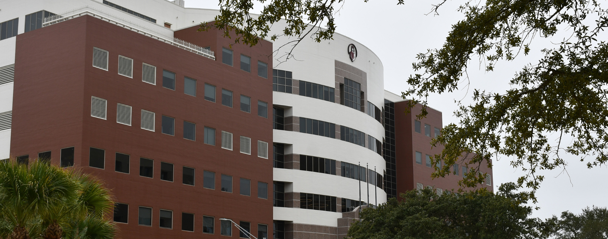 Exterior view of a multi-story UTMB Health building with red brick and white curved façade, large windows, and surrounding trees.