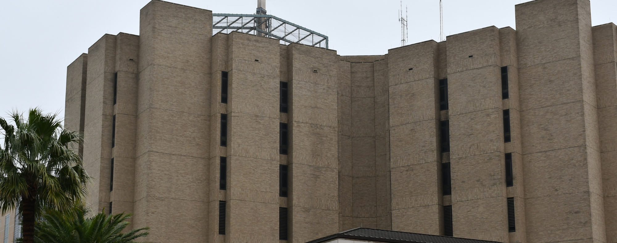 Exterior view of a large UTMB Health building with tall vertical brick sections, narrow windows, and rooftop structures framed by trees.