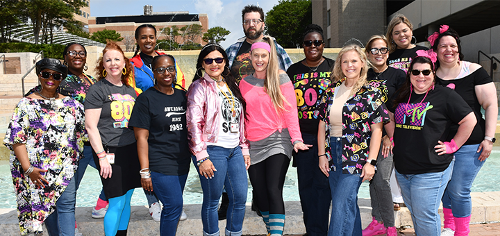 Group of individuals wearing colorful 1980s-themed outfits posing outdoors in front of a water feature and campus buildings.