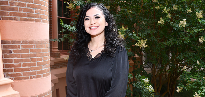 Individual with shoulder-length dark curly hair wearing a black blouse, standing outdoors near a brick building with greenery in the background.