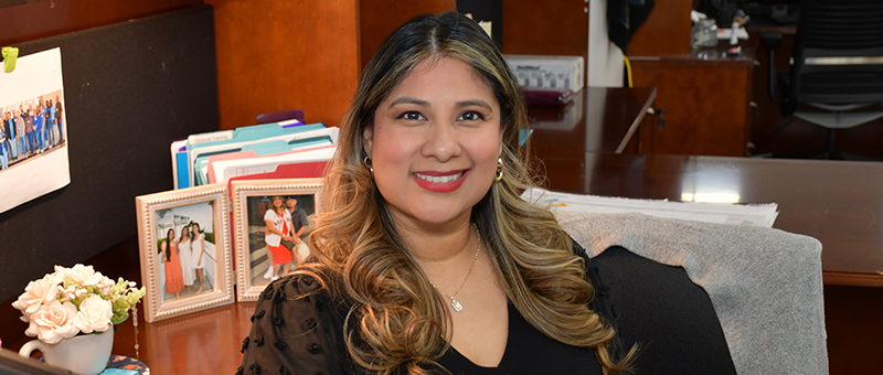 Individual seated at an office desk smiling, with framed photos, folders, and office supplies visible in the background.