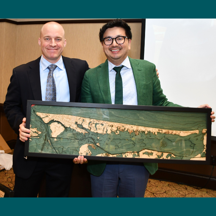 Two individuals in business attire holding a long framed map-style artwork indoors during a presentation.