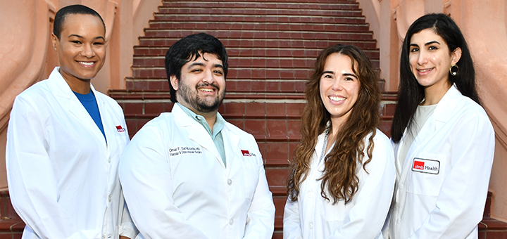 Four individuals in white medical coats stand side by side on a staircase, facing the camera in an outdoor campus setting.