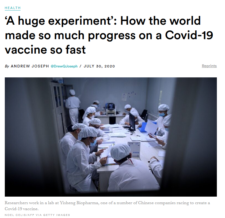 A group of laboratory researchers wearing white protective clothing, hair covers, and face masks sit around a table reviewing documents and data in a clinical lab setting during COVID-19 vaccine research.