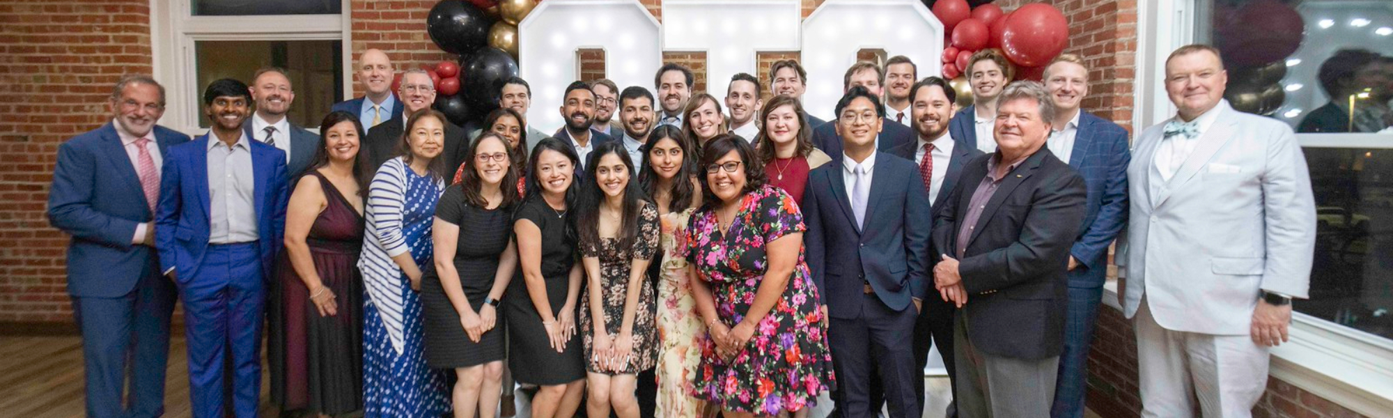 Large group of individuals standing together indoors at a formal event, with decorative balloons and illuminated letters displayed behind them.