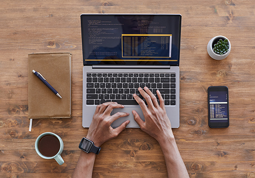 Person typing code on a laptop at a wooden desk with a notebook, phone, coffee mug, and small plant nearby.