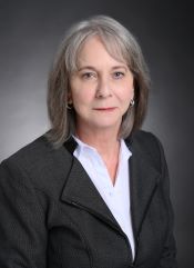 Professional headshot of Gigi Jamison with gray hair wearing a dark gray blazer and white collared shirt against a dark gray background