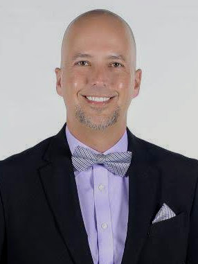 Professional headshot of Justin Robison wearing a black suit jacket, light purple dress shirt, and patterned bow tie with a matching pocket square against a white background.