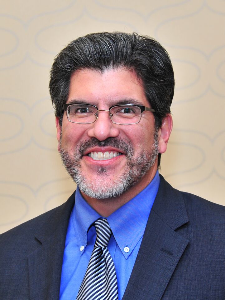 Professional headshot of Dr. Joseph Penn wearing glasses, a dark suit jacket, blue dress shirt, and striped tie against a beige patterned background.
