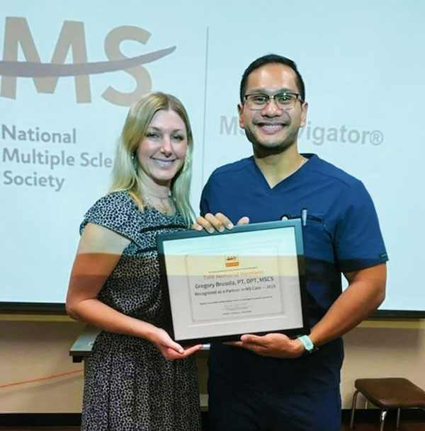 Brusola accepts an award while standing next to a woman in front of a powerpoint presentation.