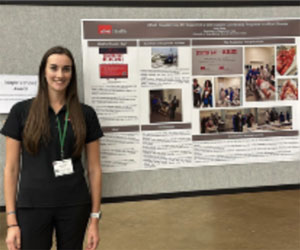 A young smiling woman stands in front of her poster presentation.