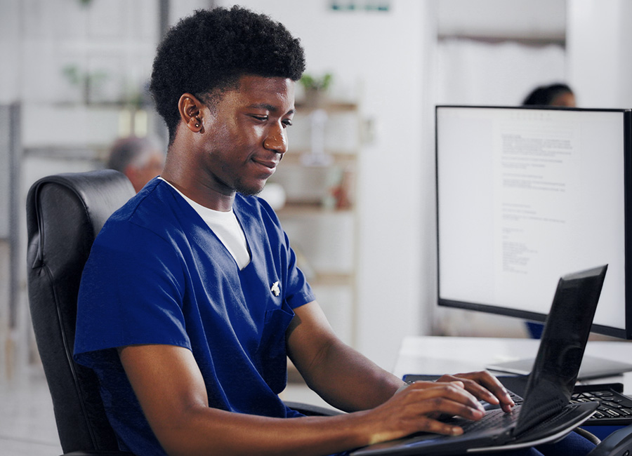 A healthcare professional in blue scrubs works on a laptop at a clinical workstation.