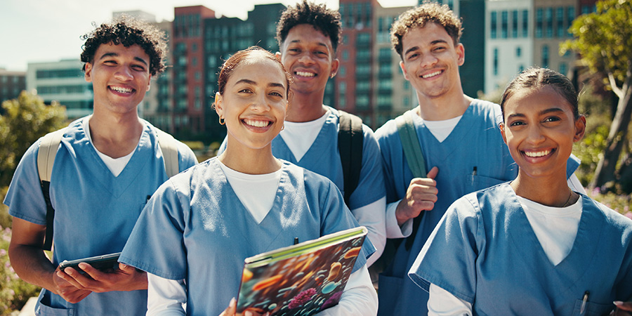 A group of healthcare students in blue scrubs stands together outside on a college campus.