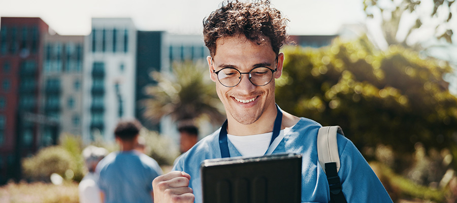 A healthcare student in blue scrubs reviews information on a tablet while standing outside on campus.