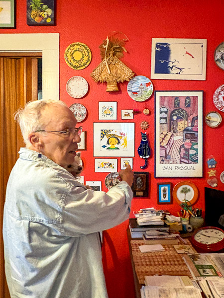 Bets Anderson, an older adult in her home pointing to a bright red wall willed with artwork