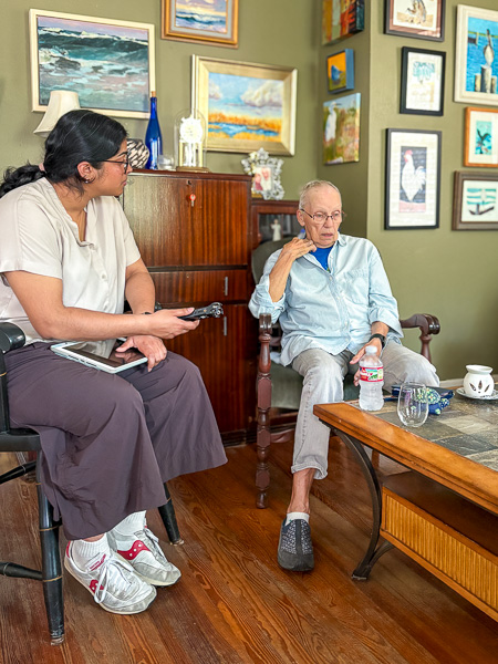 A medical student, (Annika Nambiar) interviews an older adult, (Bets Anderson) in her home holding a recording device, Bets appears thoughtful