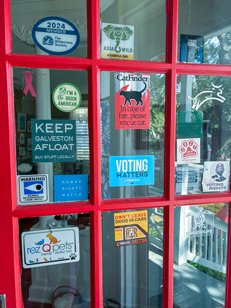Red front door with numerous stickers about various causes, “Keep Galveston Afloat”, “Voting Matters”, etc. 