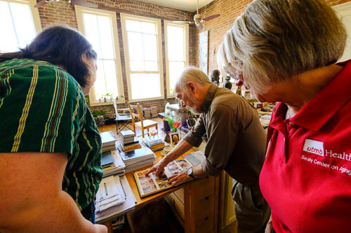 an older man points at a book as he stands talking with two women in his home