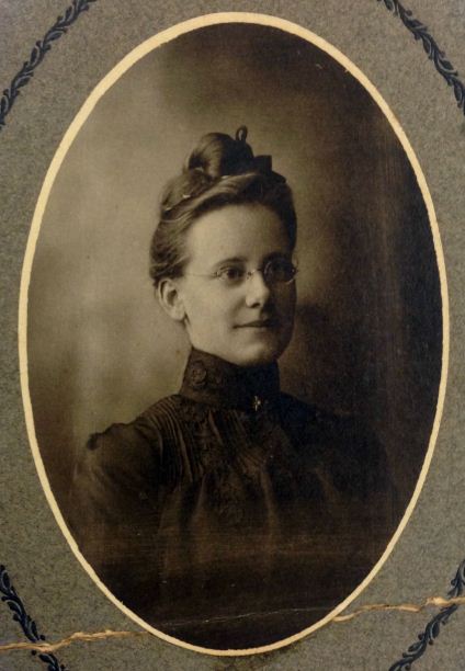 Vintage sepia tone portrait of woman, circa early 1900s. She is dress in dark high neck garment and wears glasses; her hair is in bun on top of her head.