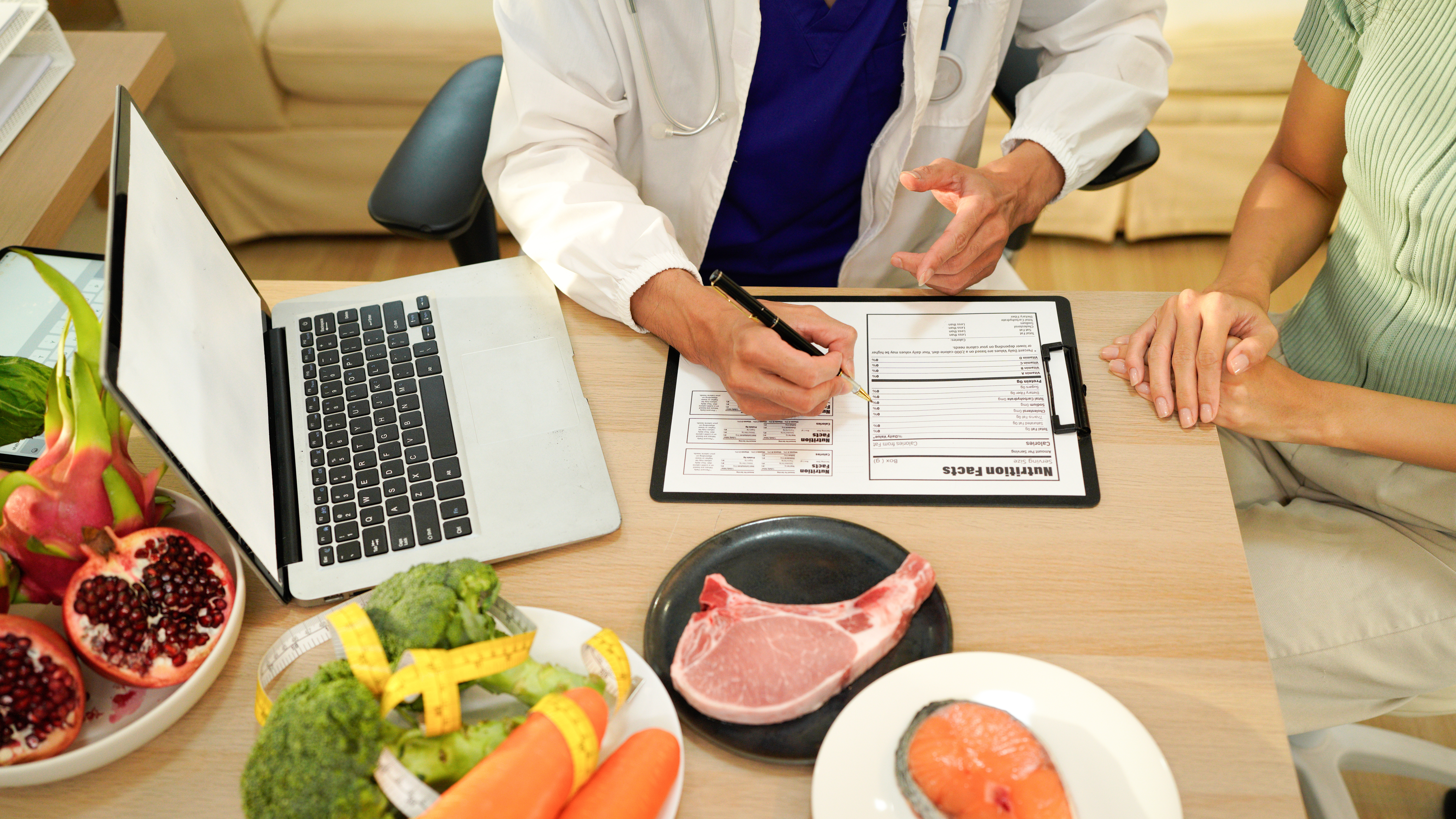 Female doctor going over nutrition facts table with another woman; a laptop, along with different foods in plates, spread out on table