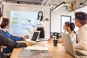 A woman is presenting to a group of people sitting around a conference table. She is pointing to call on a participant, and there is a slide showing a flowchart on the screen behind her.