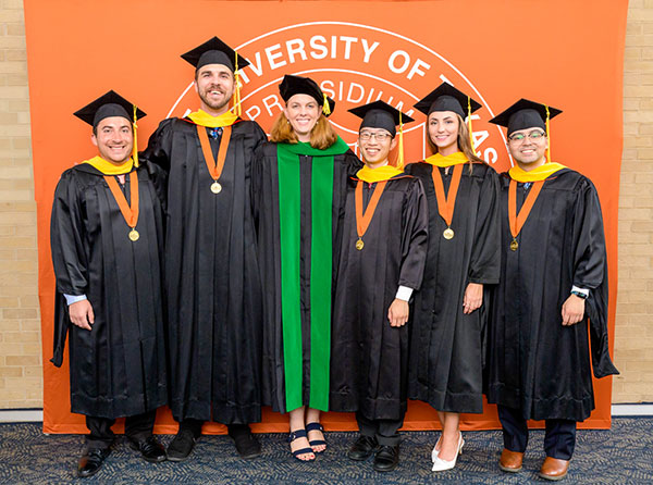 Five Aerospace Medicine graduates pose with their program director at the 2025 commencement ceremony. All are wearing caps and gowns.