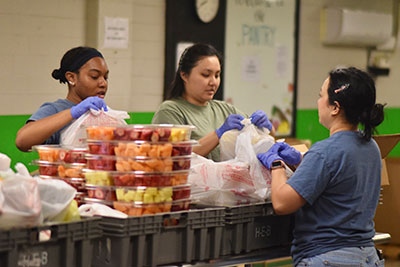 Three students are bagging up food during a food pantry volunteer event. Several plastic containers of cut fruit are seen next to them.