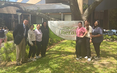 Dean David Vines and six representatives from the Occupational Therapy team hold a banner recognizing OT Month outside the SHP/SON Building.