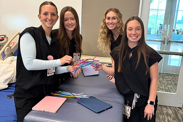 Four women in black scrubs stand on either side of a clinic bed, holding colorful wires used to teach about nerves.