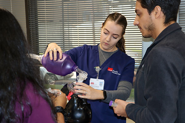 A woman with brown hair wearing UTMB Occupational Therapy scrubs is practicing intubating with a model as others watch the process.