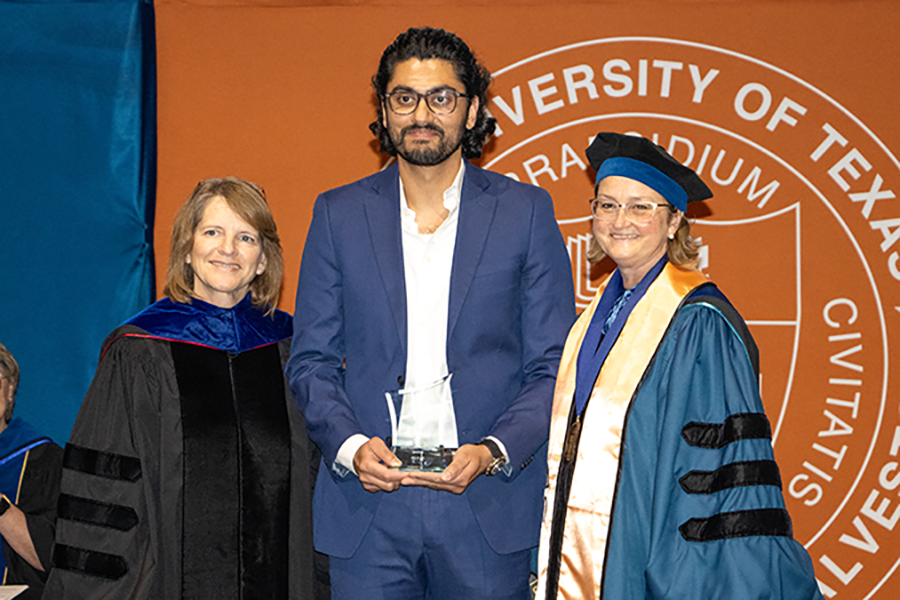Dean Melinda Sheffield-Moore and Dean Kristen Peek, both in commencement regalia, stand on either side of a man who is holding a glass award