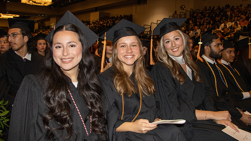 Three smiling women in caps and gowns are seated among their fellow graduates at the School of Nursing commencement ceremony.