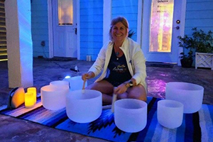 A woman instructor sits on a patio among bowls to be used as instruments in a sound bath. She is sitting on a colorful rug with lit candles off to the side.