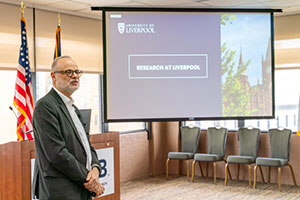 Prof. Hollander stands in front of a podium with a screen behind him displaying the title slide from his presentation, "Research at Liverpool."