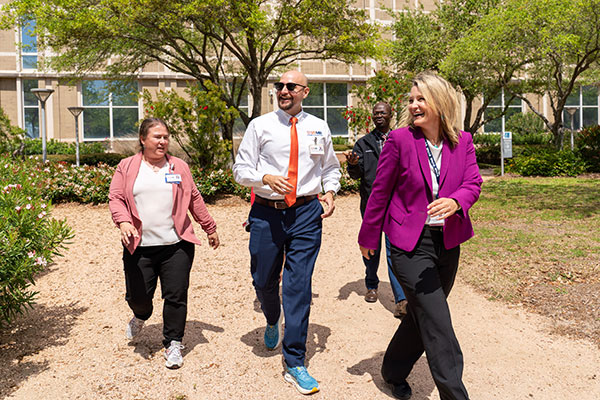 A group of colleagues take a walk outside on UTMB campus on a sunny day. The Galveston National Laboratory and several trees are in the background.